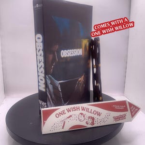 Op de afbeelding: Een zwarte VHS-cassette met de titel Obsession waarop een persoon naar een telefoon kijkt, vergezeld van een donkerbruin houten object met gouden accenten en een witte driehoekige doos met het label One Wish Willow met rode tekst.