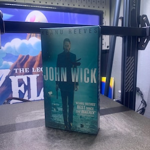 Puede incluir: Una cubierta de libro azul y verde con el título "John Wick" y el texto "Keanu Reeves' Best Since The Matrix".