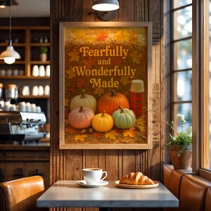 May include: A framed print with the words "Fearfully and Wonderfully Made" above a display of pumpkins and fall leaves. A red plaid thermos is next to the pumpkins. A cup of coffee and a croissant sit on a table.
