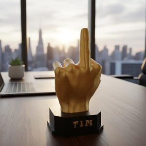 Pu&ograve; includere: Una scultura a forma di mano color oro con il dito medio alzato, montata su una base nera. La base ha il nome "TIM" in lettere dorate. La scultura &egrave; su una scrivania di legno con un laptop e una piccola pianta sullo sfondo.