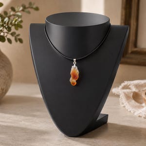 May include: A pendant necklace displayed on a black jewelry bust. The pendant is a natural stone with shades of orange and brown. The necklace cord is black, and the pendant has a silver-colored bail.
