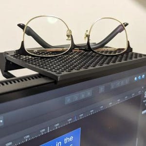 Może przedstawiać: Czarny metalowy stojak na monitor z siatką na g&oacute;rze. Na stojaku leży para okular&oacute;w w złotych oprawkach z przezroczystymi szkłami.