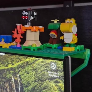 Może przedstawiać: Zielona plastikowa p&oacute;łka z r&oacute;żnymi kolorowymi figurkami Lego, w tym czarno-białą figurką z czerwonym otworem gęby, ż&oacute;łtą figurką z zielonym kapeluszem i zieloną figurką z brązowym kapeluszem. P&oacute;łka jest przymocowana do monitora komputera.