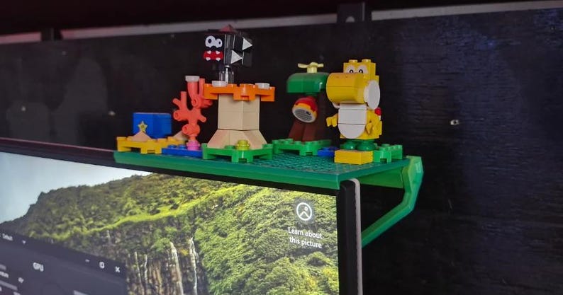 Może przedstawiać: Zielona plastikowa p&oacute;łka z r&oacute;żnymi kolorowymi figurkami Lego, w tym czarno-białą figurką z czerwonym otworem gęby, ż&oacute;łtą figurką z zielonym kapeluszem i zieloną figurką z brązowym kapeluszem. P&oacute;łka jest przymocowana do monitora komputera.