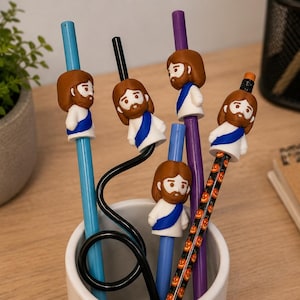 May include: A collection of pens and straws with decorative toppers featuring a cartoon depiction of Jesus. The toppers are brown, white, and blue. The pens and straws are various colors, including blue, purple, and black. The items are in a white ceramic cup.