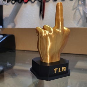 Pu&ograve; includere: Scultura a forma di mano color oro che mostra il dito medio, montata su una base esagonale nera. La base ha il nome "TIM" in lettere dorate. Scultura stampata in 3D.