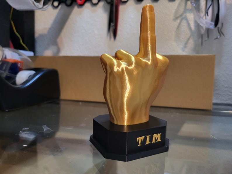 Pu&ograve; includere: Scultura a forma di mano color oro che mostra il dito medio, montata su una base esagonale nera. La base ha il nome "TIM" in lettere dorate. Scultura stampata in 3D.
