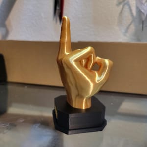 Pu&ograve; includere: Scultura a forma di mano color oro nel gesto 'ok', con l'indice rivolto verso l'alto. La mano &egrave; montata su una base geometrica nera. La scultura &egrave; probabilmente stampata in 3D e si trova su una superficie riflettente.