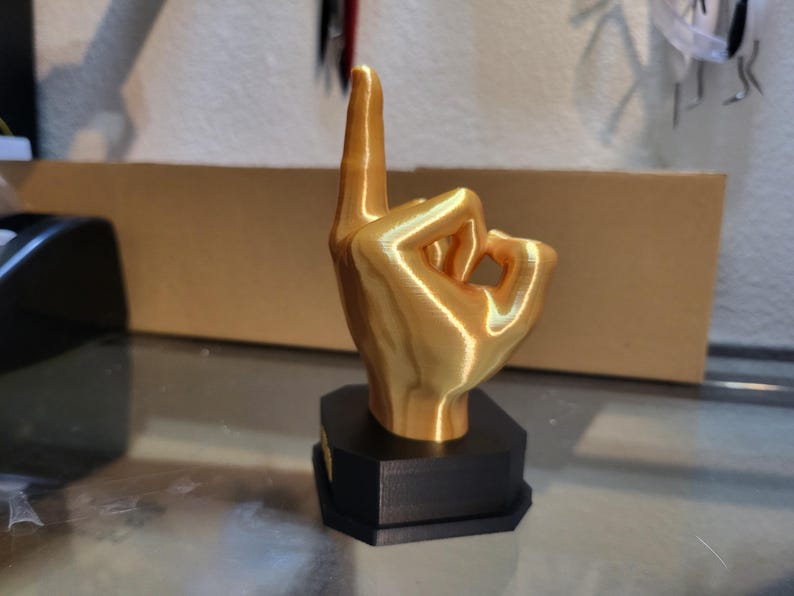 Pu&ograve; includere: Scultura a forma di mano color oro nel gesto 'ok', con l'indice rivolto verso l'alto. La mano &egrave; montata su una base geometrica nera. La scultura &egrave; probabilmente stampata in 3D e si trova su una superficie riflettente.