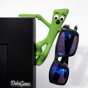 May include: A green cartoon figure, resembling a character, is holding a pair of black sunglasses with blue reflective lenses. The figure is perched on a black cabinet door. The cabinet door has a "DebsGemz" sticker.