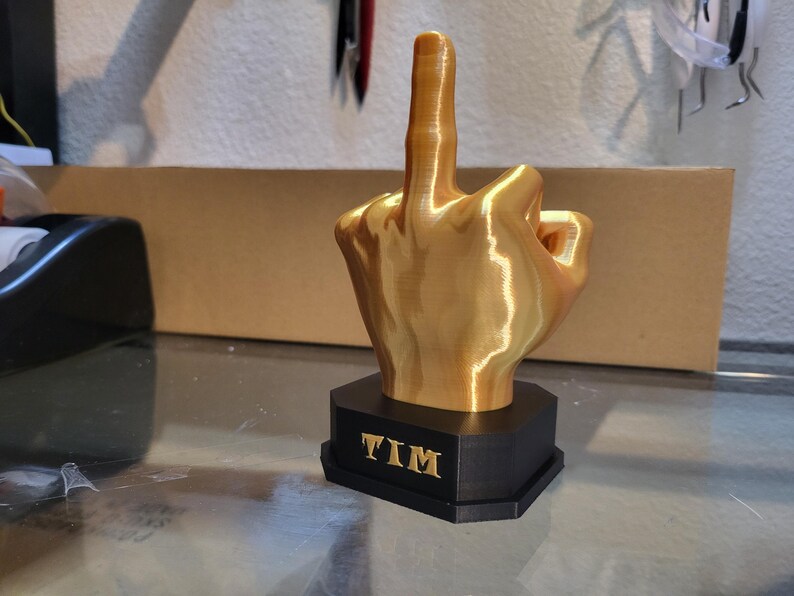 Pu&ograve; includere: Una scultura a forma di mano color oro con il dito medio rivolto verso l'alto. La scultura &egrave; montata su una base esagonale nera con il nome "TIM" in lettere dorate. La scultura &egrave; probabilmente stampata in 3D.