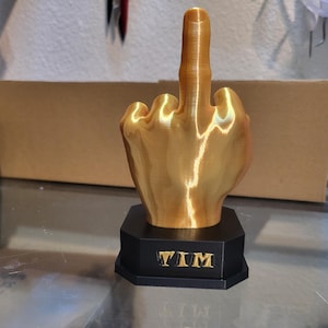Pu&ograve; includere: Una scultura a forma di mano color oro con il dito medio rivolto verso l'alto. La scultura &egrave; montata su una base nera ottagonale con il nome "TIM" in lettere dorate. La mano &egrave; molto dettagliata.