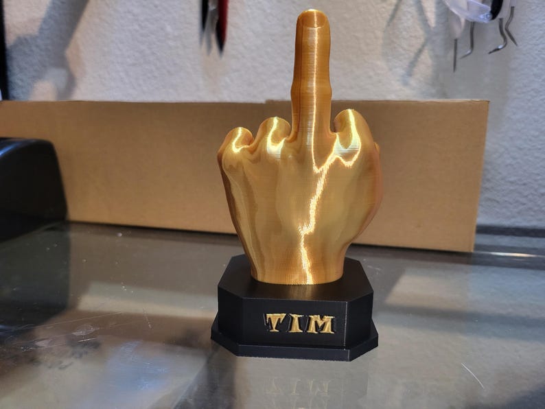 Pu&ograve; includere: Una scultura a forma di mano color oro con il dito medio rivolto verso l'alto. La scultura &egrave; montata su una base nera ottagonale con il nome "TIM" in lettere dorate. La mano &egrave; molto dettagliata.