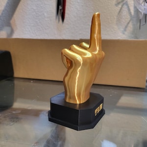 Pu&ograve; includere: Una scultura a forma di mano dorata con l'indice rivolto verso l'alto, montata su una base nera. La base presenta la scritta "WIN" in lettere dorate. La scultura &egrave; esposta su una superficie di vetro.