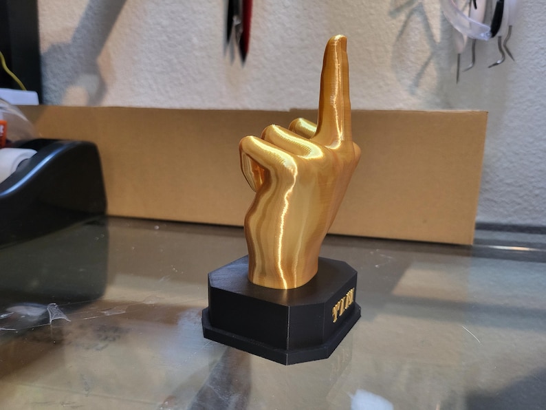 Pu&ograve; includere: Una scultura a forma di mano dorata con l'indice rivolto verso l'alto, montata su una base nera. La base presenta la scritta "WIN" in lettere dorate. La scultura &egrave; esposta su una superficie di vetro.