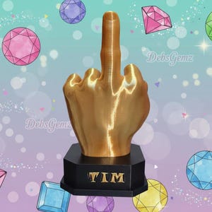 Pu&ograve; includere: Una scultura a forma di mano color oro che mostra il dito medio, montata su una base nera con il nome "TIM". Lo sfondo presenta gemme colorate e il testo "DebsGemz".