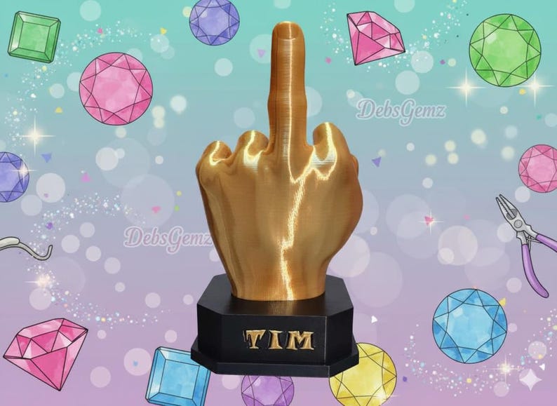 Pu&ograve; includere: Una scultura a forma di mano color oro che mostra il dito medio, montata su una base nera con il nome "TIM". Lo sfondo presenta gemme colorate e il testo "DebsGemz".