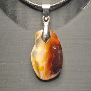 May include: A pendant necklace featuring a polished, oval-shaped stone with a silver-tone bail. The stone displays a mix of colors, including orange, brown, and cream. The necklace has a black cord.