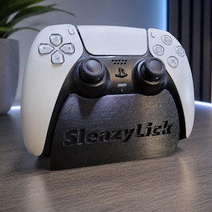 May include: A white and black wireless game controller resting on a black stand. The stand features the text SleazyLick embossed on the front. The controller has two black analog sticks, a central touchpad, and standard directional and action buttons.