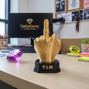 May include: A gold-colored hand sculpture giving the middle finger, mounted on a black base with the name "TIM" in gold lettering. The sculpture is on a desk with a computer and other office supplies.
