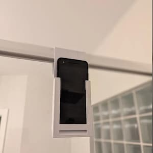 Puede incluir: Soporte para teléfono de plástico blanco fijado a una puerta de ducha de vidrio. El soporte tiene una ranura para un teléfono y un borde para evitar que el teléfono se caiga.