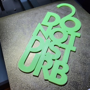 Può includere: Un cartello verde brillante stampato in 3D con la scritta "DO NOT DISTURB". Le lettere sono sovrapposte, creando un design giocoso e accattivante. Il cartello è probabilmente per una porta o una scrivania.