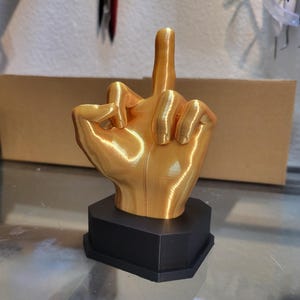 Pu&ograve; includere: Una scultura a forma di mano color oro con il dito medio rivolto verso l'alto, montata su una base esagonale nera. La mano &egrave; dettagliata con linee e texture visibili, suggerendo un design stampato in 3D. La scultura &egrave; esposta su una superficie riflettente.