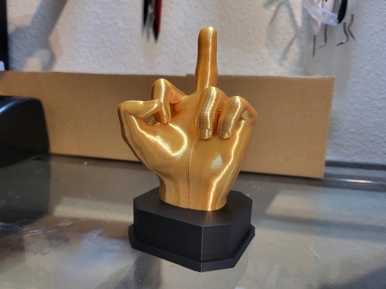 Pu&ograve; includere: Una scultura a forma di mano color oro con il dito medio rivolto verso l'alto, montata su una base esagonale nera. La mano &egrave; dettagliata con linee e texture visibili, suggerendo un design stampato in 3D. La scultura &egrave; esposta su una superficie riflettente.
