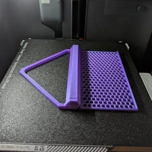 Może przedstawiać: Fioletowy przedmiot wydrukowany w 3D z wzorem plastra miodu i tr&oacute;jkątnym uchwytem. Przedmiot został zaprojektowany do użycia jako stojak na telefon.