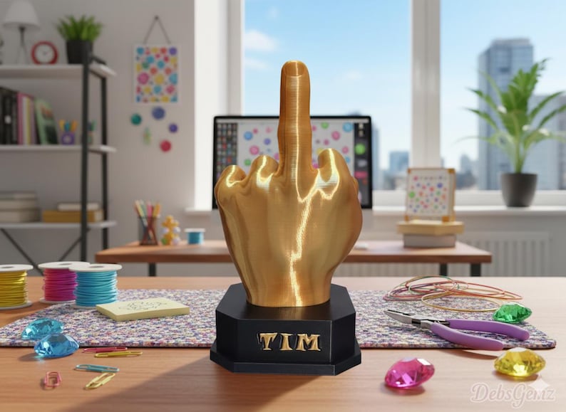 Pu&ograve; includere: Una scultura a forma di mano color oro che mostra il dito medio, montata su una base nera con il nome "TIM" in lettere dorate. La scultura &egrave; su una scrivania con materiali per l'artigianato e un computer sullo sfondo.