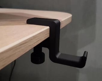 May include: A black metal headphone hanger clamped to the edge of a light-coloured wooden desk. The hanger has a curved design with a hook to hold headphones and a screw to secure it to the desk.