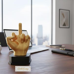 Pu&ograve; includere: Una scultura a forma di mano color oro con il dito medio alzato, montata su una base nera. La scultura &egrave; su una scrivania di legno con un laptop, un taccuino, una penna e una piccola pianta grassa in un vaso bianco. Lo sfondo include una finestra con vista sulla citt&agrave;.