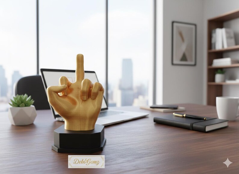 Pu&ograve; includere: Una scultura a forma di mano color oro con il dito medio alzato, montata su una base nera. La scultura &egrave; su una scrivania di legno con un laptop, un taccuino, una penna e una piccola pianta grassa in un vaso bianco. Lo sfondo include una finestra con vista sulla citt&agrave;.