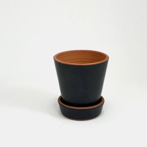 May include: A black terracotta planter with a matching saucer. The planter has a rounded bottom and a slightly flared top. The saucer is flat and has a slightly raised edge.