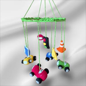May include: A handmade crochet mobile with a green ring and hanging elements. The mobile features colourful crochet cars in red, yellow, blue, and green, plus a traffic light, a road cone, and a banana. The mobile is suspended by green strings.