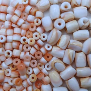 May include: A collection of small, cylindrical beads in two distinct sizes and colors. The smaller beads are a soft peach-orange hue, while the larger beads are a creamy white with subtle, natural-looking tan and light orange color variations.