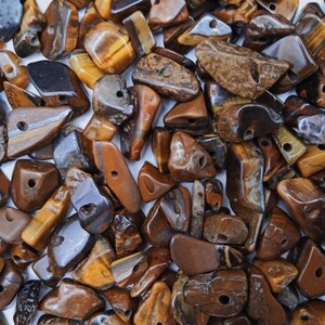 May include: A collection of loose, tumbled tiger's eye gemstone beads in various irregular shapes and sizes. Each stone features a polished, glossy finish with characteristic golden-brown, dark brown, and black bands, and includes a drilled hole.
