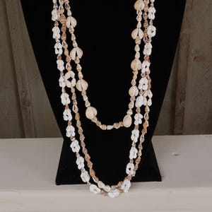 May include: A long multi-strand necklace composed of various small white and tan seashells. The necklace features a layered design with alternating patterns of light-colored shells strung together to create a continuous, textured, and organic appearance.