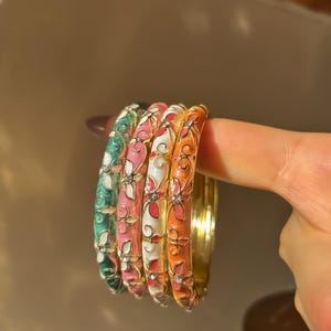 May include: A set of enamel bangles in various colours. Each bangle has a floral pattern in green, pink, white, and orange, with small, sparkling details. The bangles are gold-coloured and are being held up.