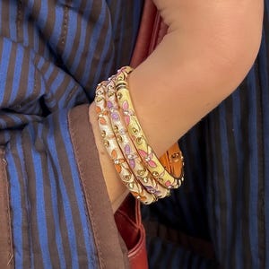 May include: Four gold bangles with floral designs in pink, orange, and purple. The bangles are worn on an arm, and the background includes a blue and brown striped pattern.
