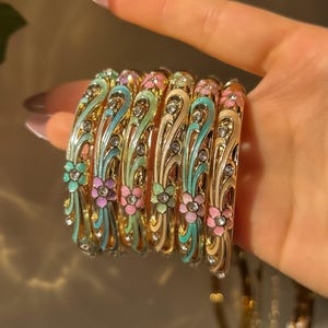 May include: Gold-toned bangles with colorful floral and scroll designs. Each bangle features a different color scheme, including pink, blue, and green enamel, accented with clear crystals. The bracelets are ornate and elegant.