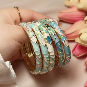 Könnte beinhalten: Eine Sammlung goldfarbener Armreifen mit aufwendigen Emaille-Designs. Die Armreifen haben eine hellblaue Basis mit weißen, rosa und grünen Blumenmustern. Die Armbänder sind gestapelt, was ihre detaillierte Handwerkskunst zeigt. Jeder Armreif hat einen Durchmesser von ca. 6 cm.