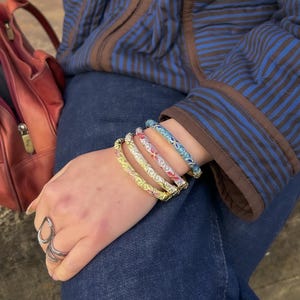 Può includere: Una collezione di braccialetti colorati con motivi floreali. I braccialetti sono gialli, rosa, rossi e blu. La persona indossa una giacca blu e jeans.