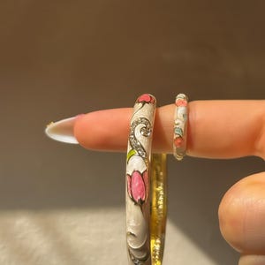 May include: A cream-colored bangle bracelet and a matching ring, both featuring an enamel floral design with pink and green accents, gold-tone metal edges, and small embedded clear crystals arranged in swirling patterns along the surface.