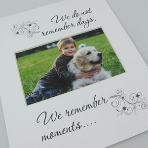 May include: A white photo frame with a black and white floral design on the top and bottom. The frame has a photo of a young boy hugging a golden retriever dog. The text "We do not remember days, We remember moments..." is printed on the frame.