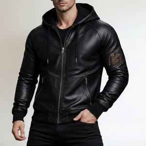 Può includere: Giacca con cappuccio in pelle nera con chiusura a zip, tasche frontali e orlo e polsini a coste. La giacca presenta un emblema decorativo sulla manica superiore sinistra. Il modello indossa pantaloni neri.
