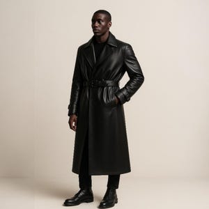 May include: A full-length black leather trench coat with a belted waist and notched lapels. The coat is worn with black pants and black boots. The model is standing in front of a neutral background.