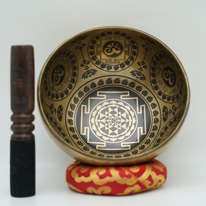 May include: A gold singing bowl with a black and white geometric design, a wooden striker, and a red cushion.