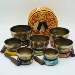 May include: Set of seven singing bowls with wooden strikers and colorful cushions. The bowls are made of brass and have intricate designs. The bowls are arranged in a pyramid shape with the largest bowl at the bottom and the smallest bowl at the top.