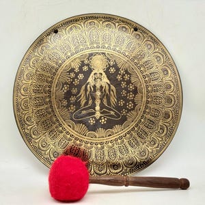 Puede incluir: Un gong grande, redondo, de latón con un diseño de mandala detallado en oro. El gong presenta una imagen central de una figura sentada con un patrón floral. Se incluye un mazo de fieltro rojo.
